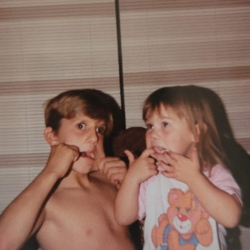 A brother and sister with light brown hair making silly faces at a kitchen table.