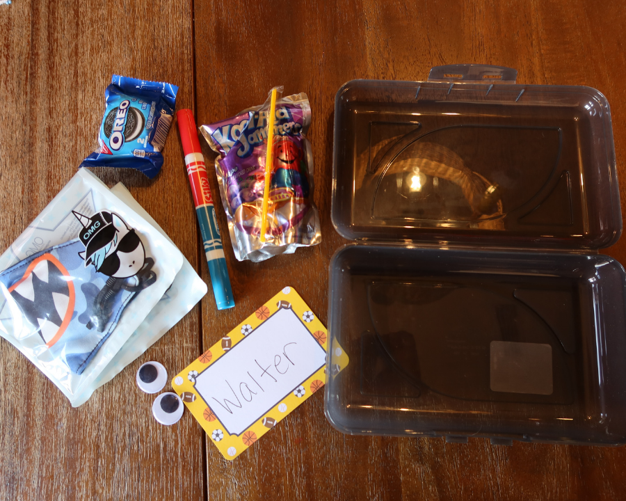 A translucent black pencil box on top of a wooden table with a blue shark mask, orange and blue scented marker, pack of oreos, grape capri sun, name tag and a pair of googly eyes laying beside it.