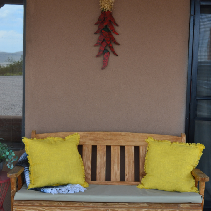 image of a weathered bench behind a stucco wall with a chile ristra. The bench has a tan cushion with two yello pillows and a blue tassel blanket.