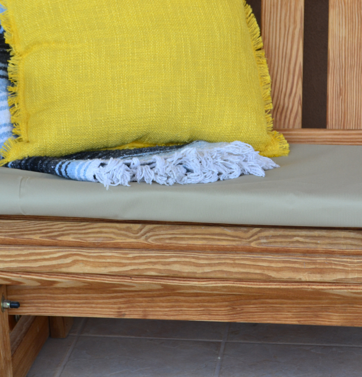 close up image of a weathered wooden bench and a beige cushion with a yellow pillow and blue tassel blanket